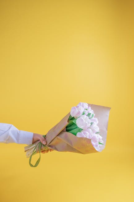 A person wearing a white shirt is holding a bouquet of fresh, light pink tulips wrapped in beige paper with a green ribbon against a vibrant yellow background. The tulips have smooth petals with some 