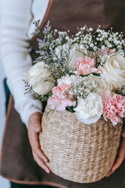 A person holding a woven wicker basket filled with a fresh floral arrangement featuring white roses, pink carnations, baby's breath, and sprigs of lavender. The flowers are vibrant and well-hydrated, 
