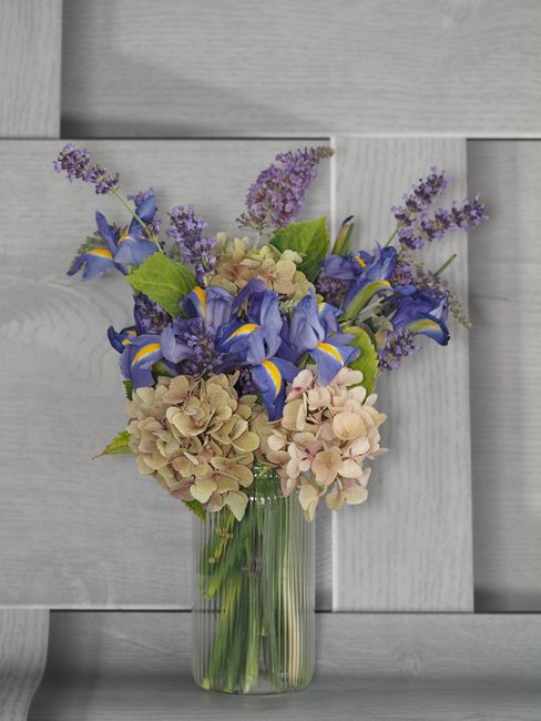 A tall, clear glass vase filled with a floral arrangement featuring soft pink hydrangeas, vibrant blue irises with yellow accents, and sprigs of purple lavender. The flowers are fresh, with lush petal