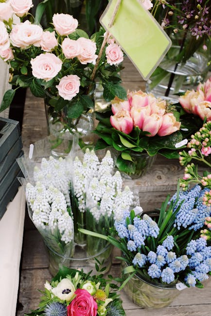 A variety of fresh flowers arranged in bouquets and vases on a wooden surface. There are pale pink roses with lush green leaves in a clear glass vase, complemented by pink and cream tulips with green