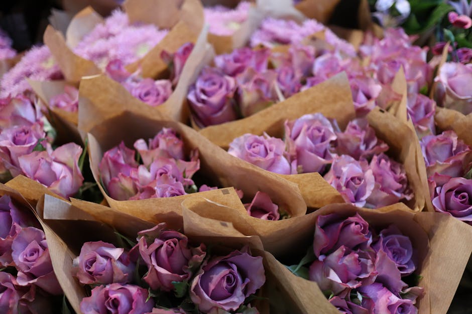 A collection of fresh lavender-pink roses, each with tightly packed petals displaying a gentle gradient from soft pink to lavender, arranged in small clusters wrapped in light brown kraft paper. The r